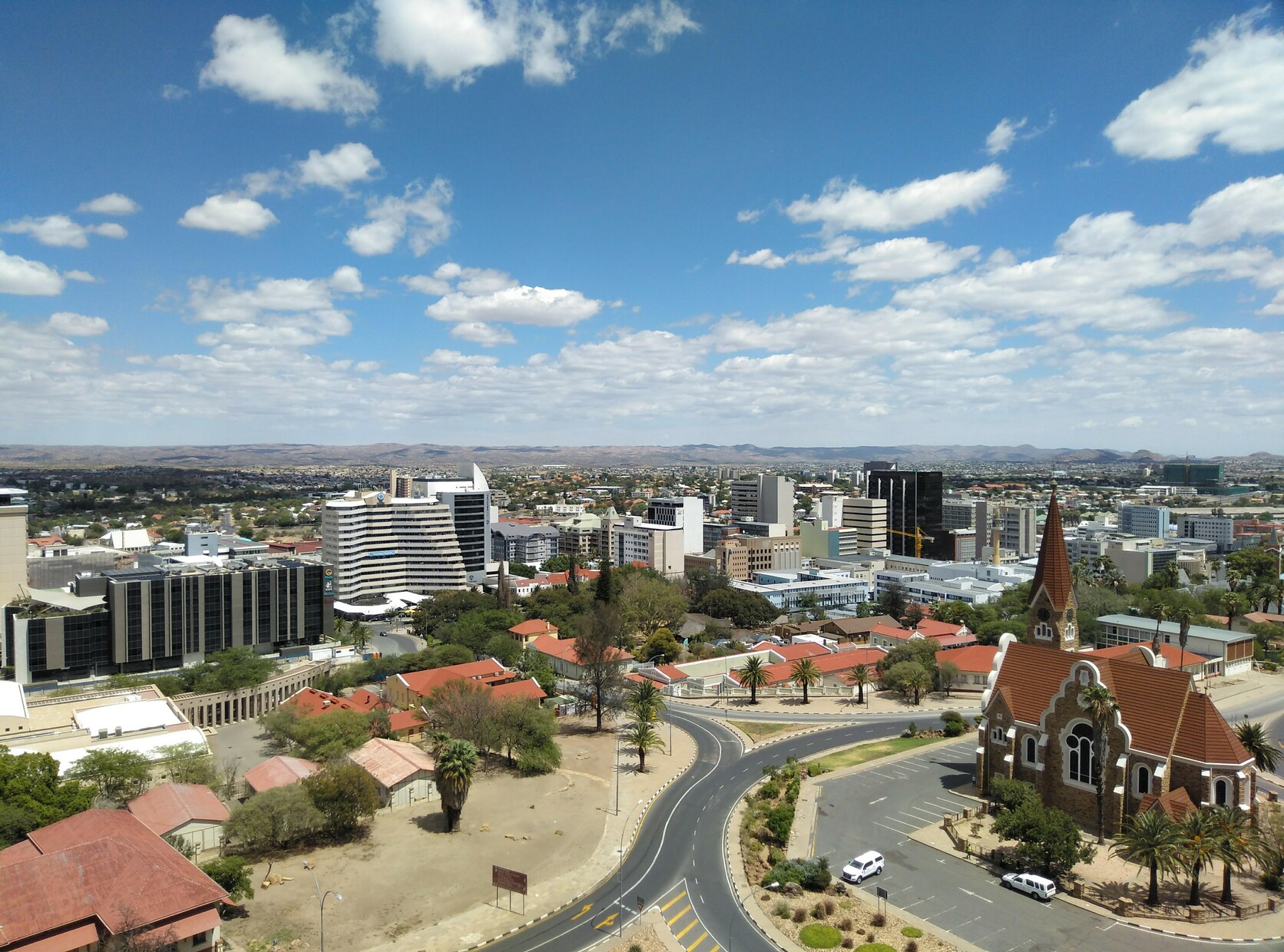 Panoramic photo of Windhoek, Namibia, showing a German-style church, a sunny sky with some clouds, some tall buildings, and the mountains in the background