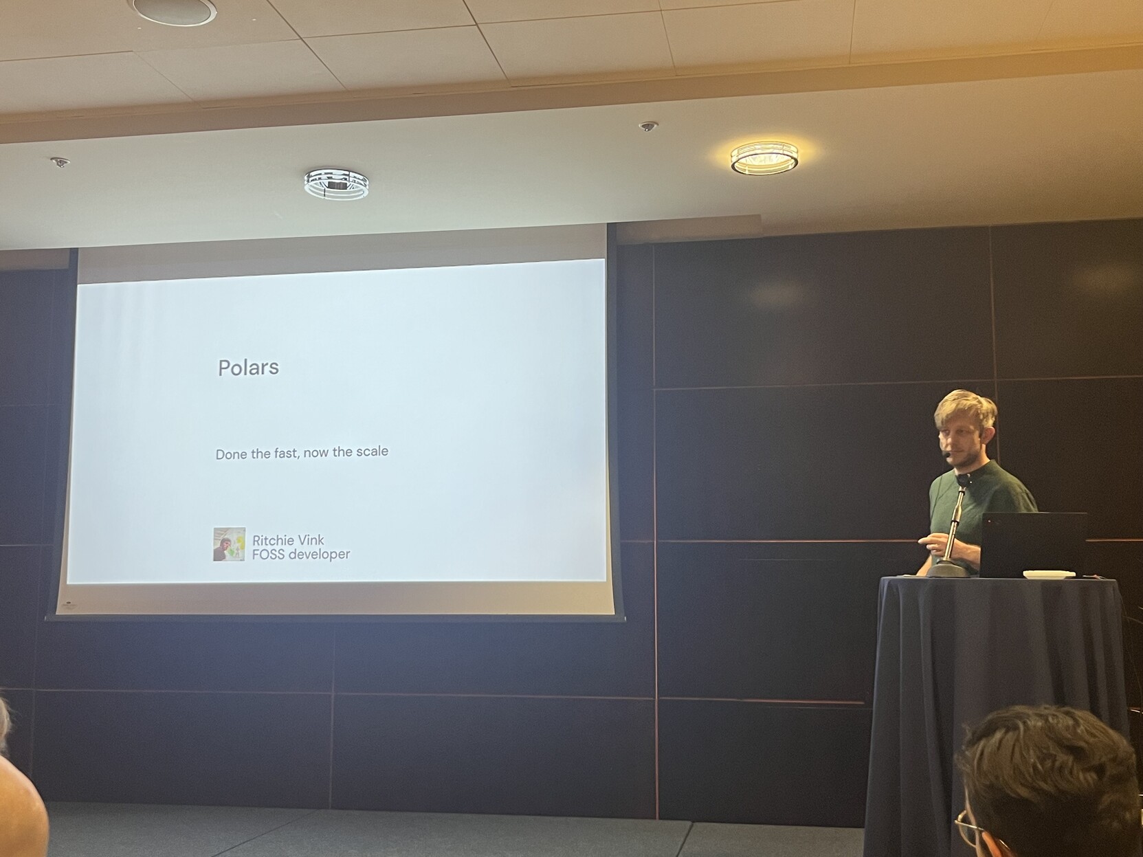 Photo of Ritchie standing next to a screen with a slide saying “Polars: Done the fast, now the scale”