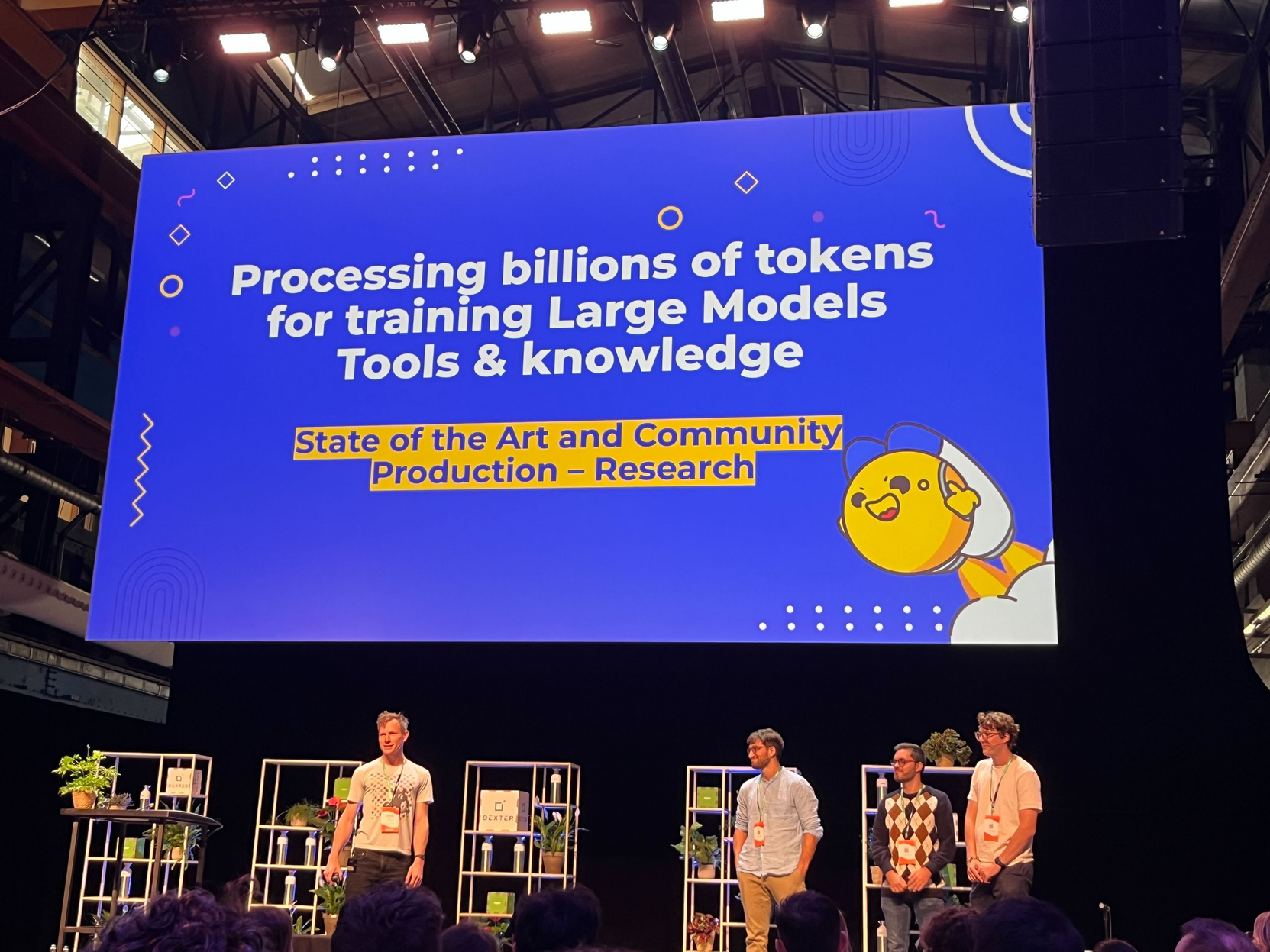 Photo of speakers under a giant screen with a slide titled "Processing billions of tokens for training Large Models Tools & knowledge"