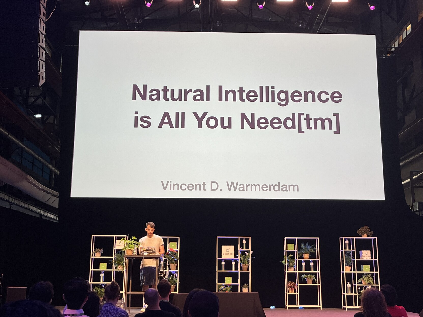 Photo of man under a giant screen, showing a slide titled "Natural Intelligence is All You Need[tm]"