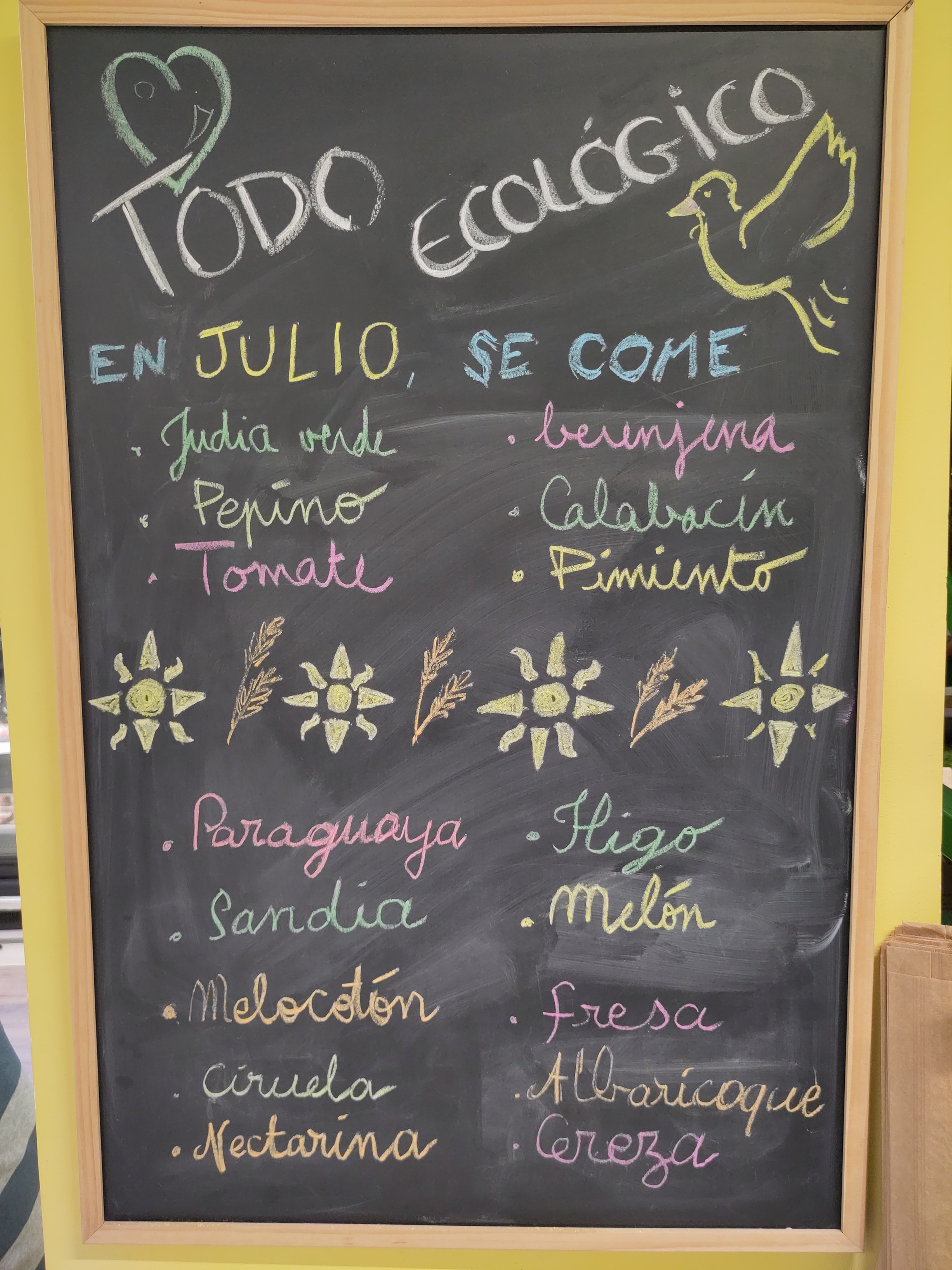 Foto de una pizarra negra pintada con tizas de varios colores, pone "Todo ecológico" y las frutas y verduras que se comen en julio